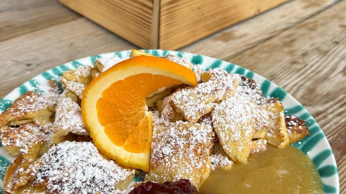 Ein Teller mit goldenen, süßen Kaiserschmarrn, bestreut mit Puderzucker und frischen Orangenscheiben. Dazu gibt es Apfelmus und Himbeermarmelade, angerichtet auf einem bunten Teller. | © Schladminger Alm