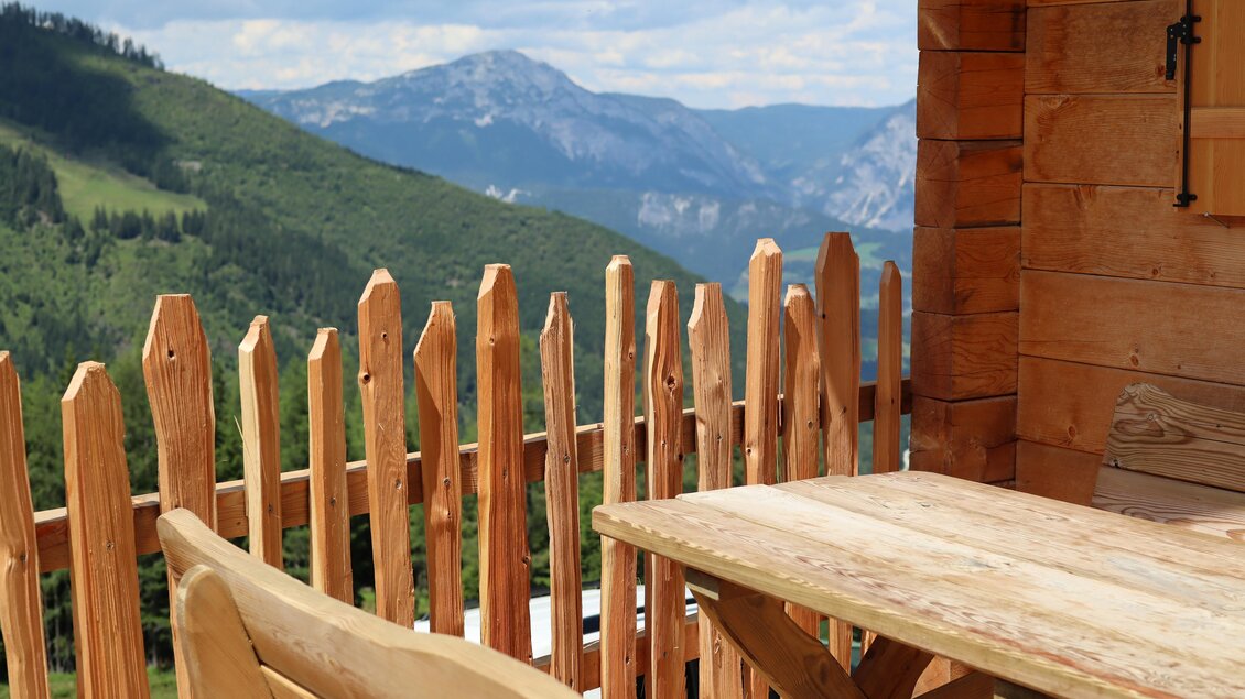 Eine gemütliche Terrasse mit einem Holztisch und einer Holzbanane, vor einer beeindruckenden Berglandschaft. Im Hintergrund sind grüne Hügel und Wolken am blauen Himmel zu sehen. | © Schladminger Alm