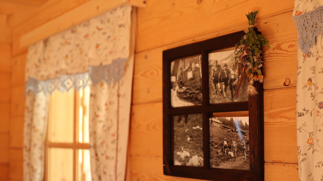 Ein rustikales Zimmer mit Holzverkleidung und blumigen Vorhängen. An der Wand hängt ein gerahmtes Bild mit historischen Fotografien. | © Schladminger Alm