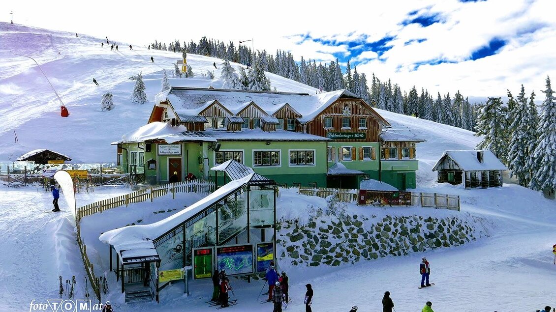 Eine schöne Berghütte im Schnee, umgeben von winterlicher Landschaft. Skifahrer und Snowboarder genießen die Pisten vor der charmanten Unterkunft. | © Foto TOM