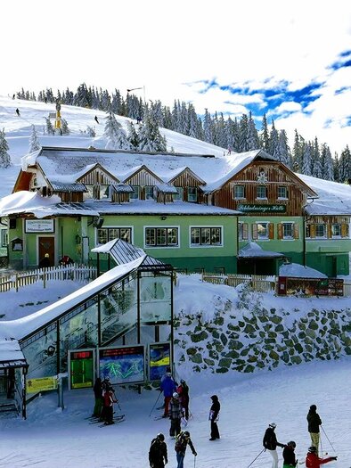 A beautiful mountain cabin in the snow, surrounded by a wintry landscape. Skiers and snowboarders enjoy the slopes in front of the charming accommodation. | © Foto TOM