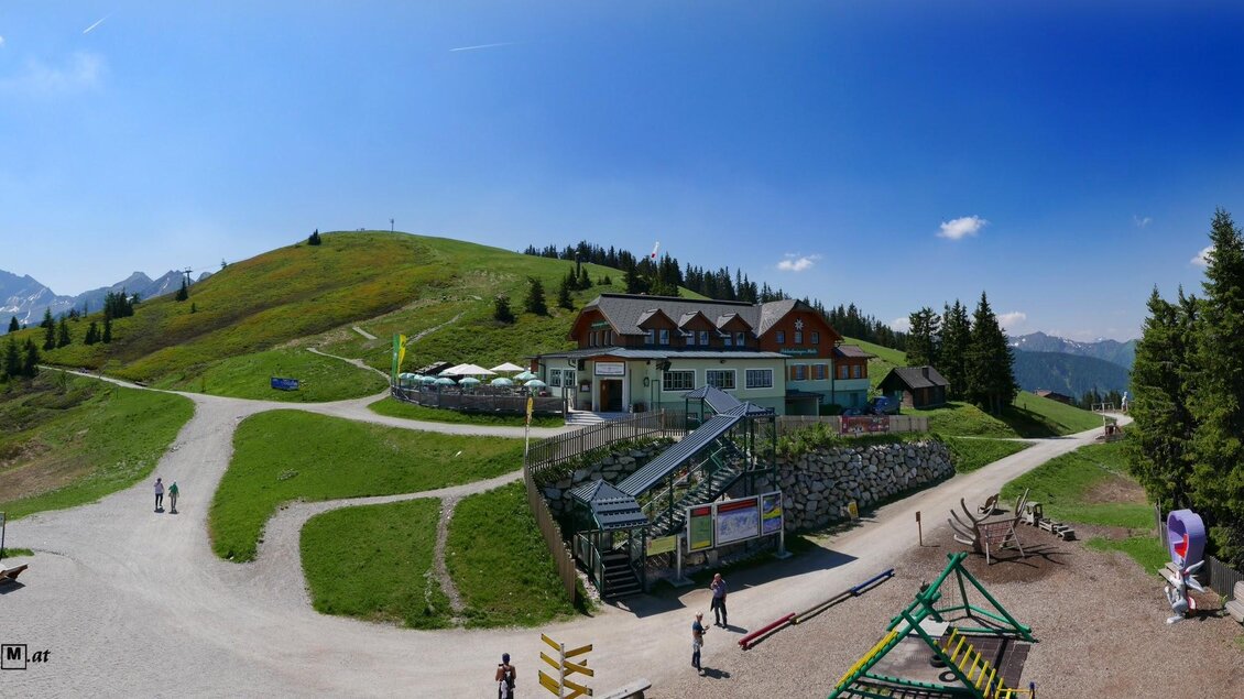 Eine grüne Berglandschaft mit einem Gebäude und mehreren Wegen. Im Vordergrund sind Spielgeräte und einige Besucher zu sehen. | © Foto TOM