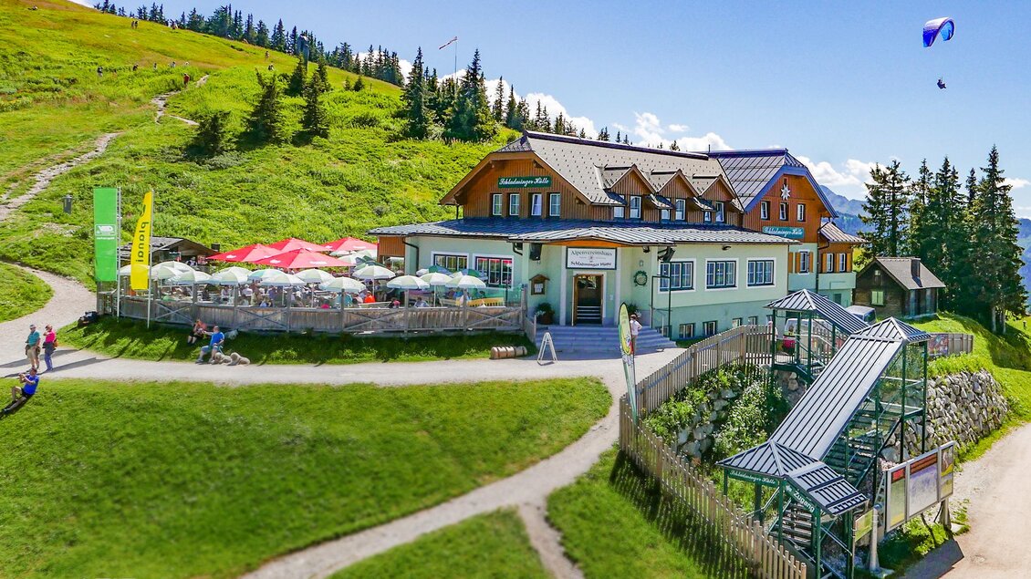 Ein malerisches Restaurant in den Bergen, umgeben von grünen Wiesen und Bäumen. Paragliding und eine Terrasse mit Sonnenschirmen laden zum Verweilen ein. | © Foto TOM