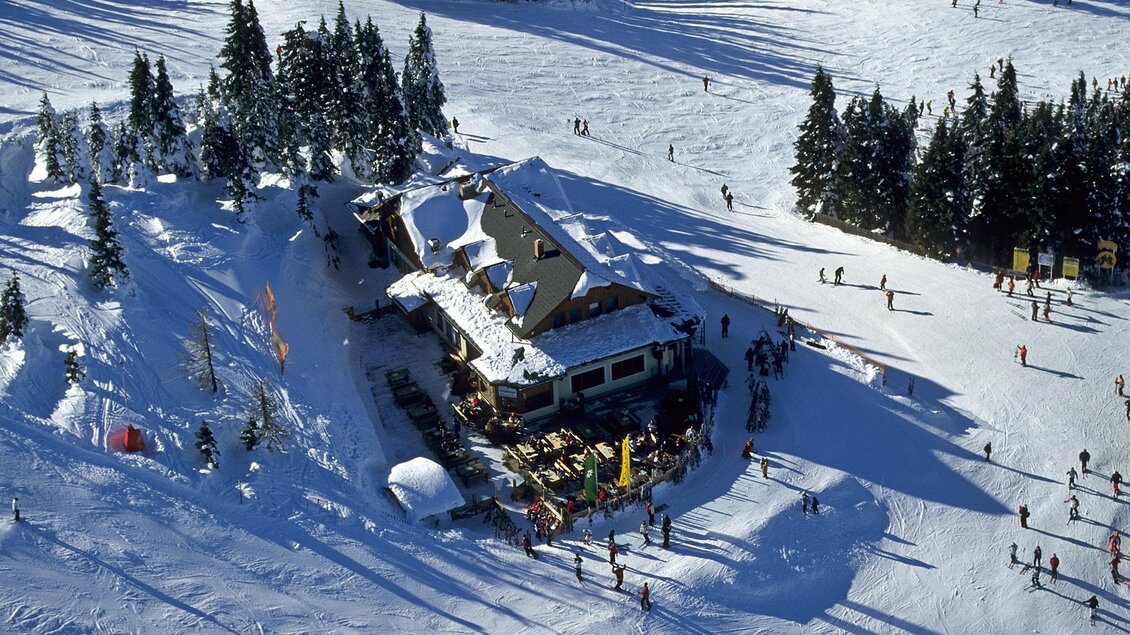 Eine gemütliche Skihütte umgeben von schneebedeckten Bäumen. Im Vordergrund sind Skifahrer und Besucher, die die Winterlandschaft genießen. | © Foto TOM