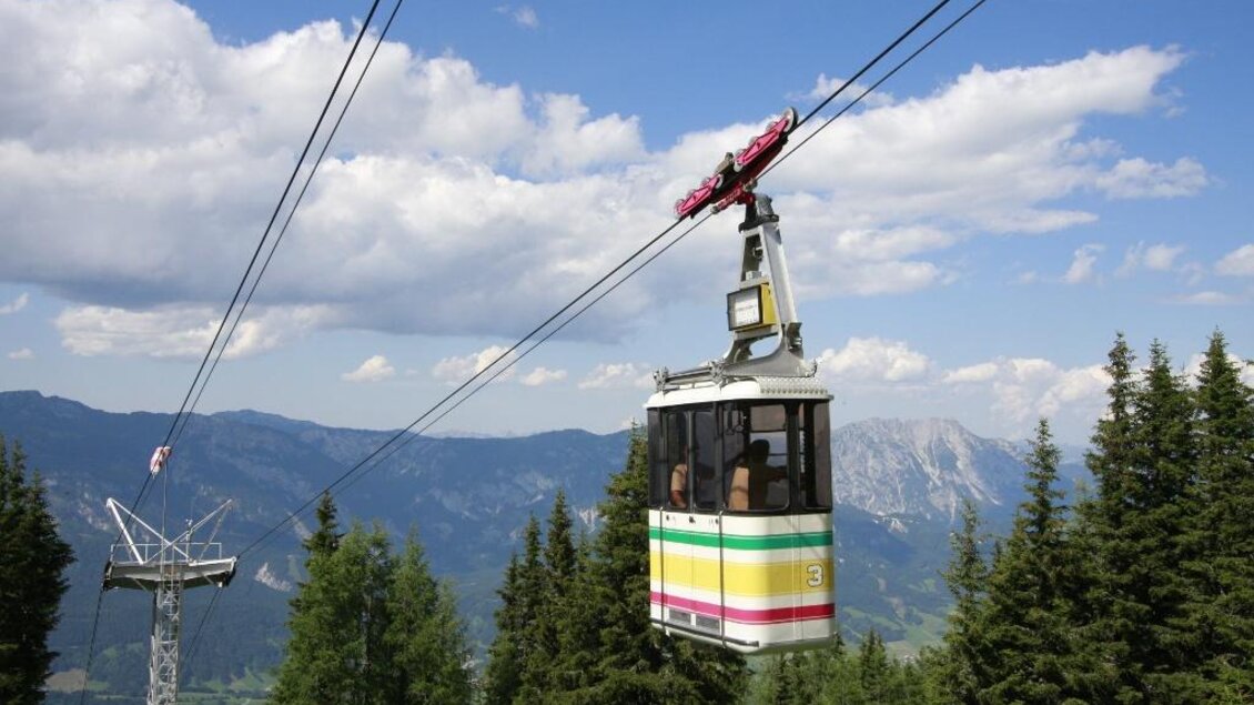 Eine Seilbahn schwebt über grüne Bäume und Berge unter einem blauen Himmel. Die umliegende Landschaft ist malerisch und einladend. | © Verein Schaferlebnis am Hauser Kaibling