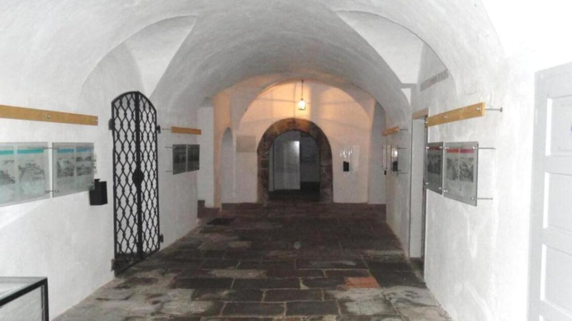 Ein langer, heller Steingang mit gewölbten Decken. An den Wänden hängen Informationen und Bilder, während eine Tür zu einem anderen Raum führt. | © NMS Köflach