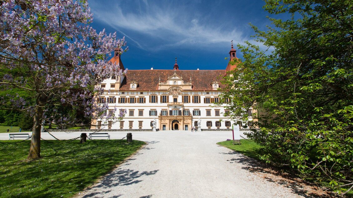 Ein beeindruckendes Schloss mit langen Gängen und roten Dächern. Vor dem Schloss befinden sich blühende Bäume und ein grüner Park. | © Graz Tourismus - Harry Schiffer