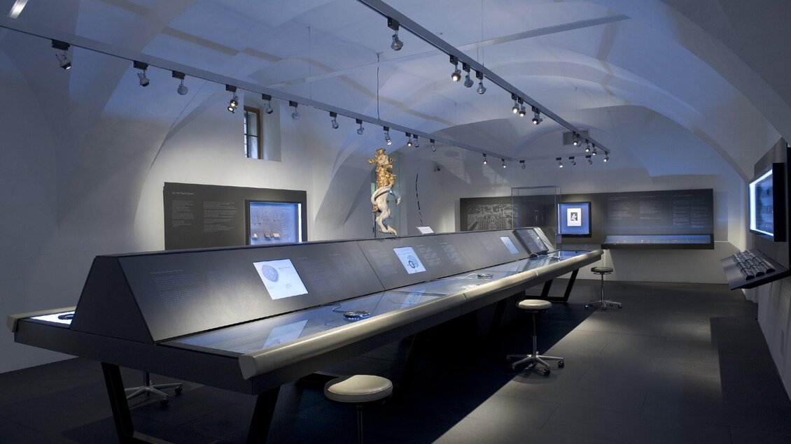 Ein modernes Ausstellungsraum mit langen Tischen und digitalen Displays. An der Decke hängt eine anatomische Skulptur. | © UMJ - Nicolas Lackner