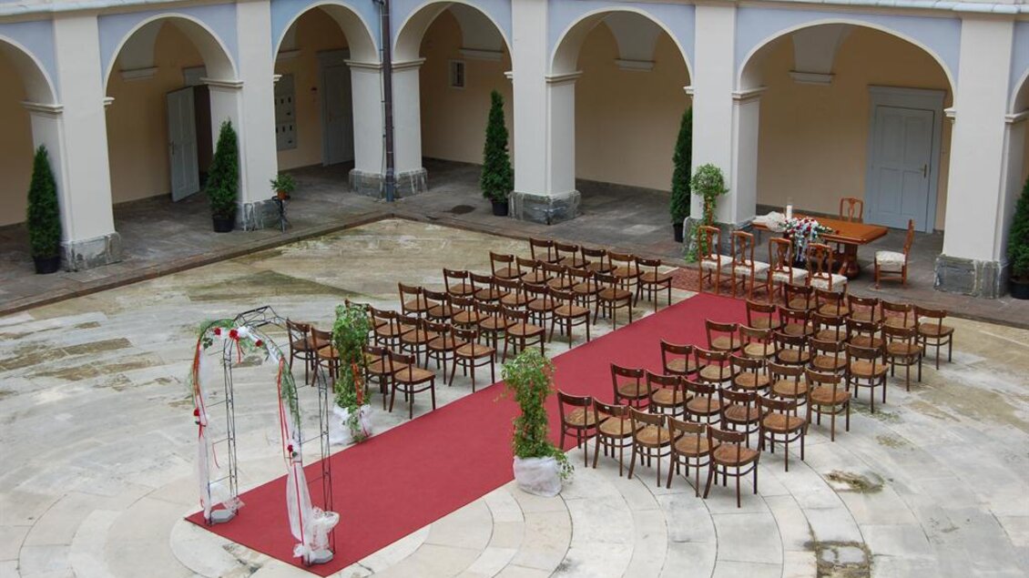 Eine elegante Hochzeitszeremonie im Innenhof mit Stühlen in Reihen und einem dekorierten Trauungsbogen. Der Boden ist mit einem roten Teppich ausgelegt und die Umgebung ist freundlich gestaltet. | © www.piber.com