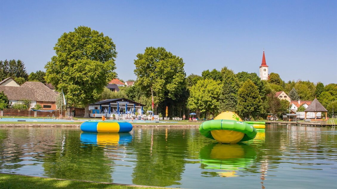 Ein ruhiger See mit bunten Wasserattraktionen schwimmt auf der Oberfläche. Im Hintergrund sieht man schattige Bäume und ein Gebäude mit einem Kirchturm. | © Sandra Brünner