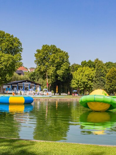 A tranquil lake with colorful water attractions floats on the surface. In the background, you can see shady trees and a building with a church tower. | © Sandra Brünner