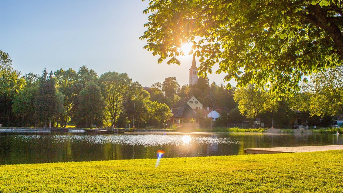 Eine ruhige Landschaft mit einem See und einer Kirche im Hintergrund. Die Sonne scheint durch die Bäume und sorgt für eine angenehme Atmosphäre. | © Sandra Brünner