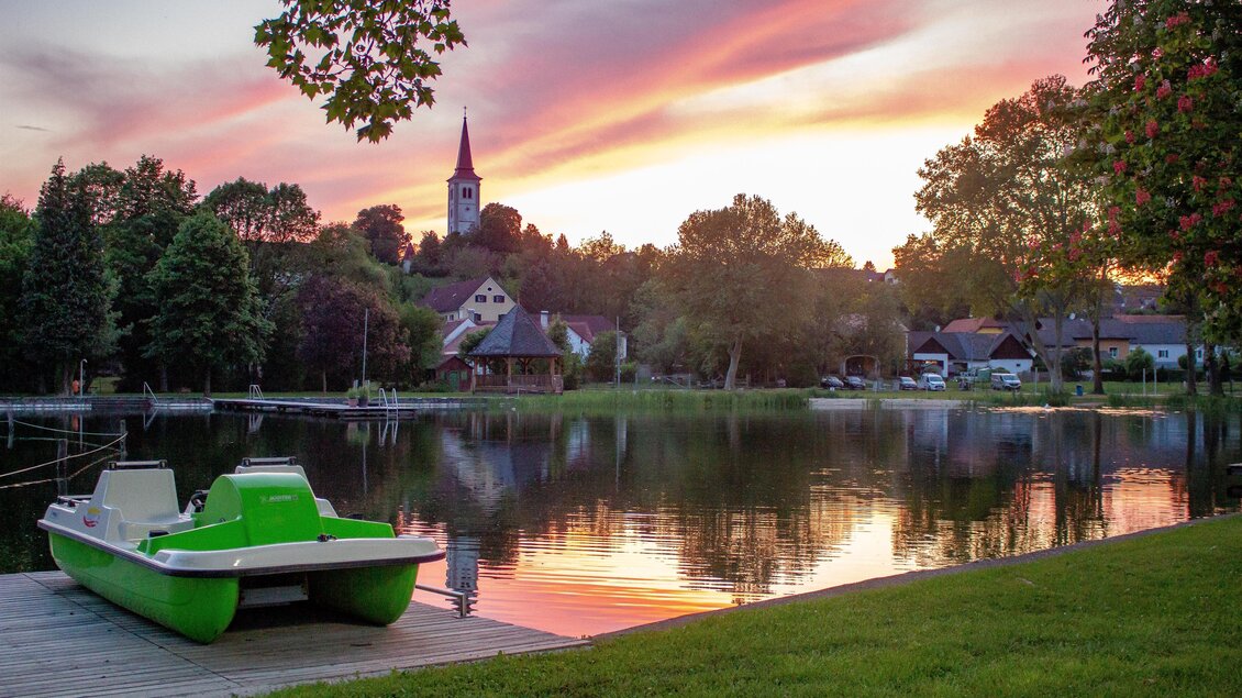 Ein ruhiger See mit einem grünen Tretboot am Ufer. Der Himmel ist in schönen Farben gehüllt, während die Sonne untergeht. | © Sandra Brünner