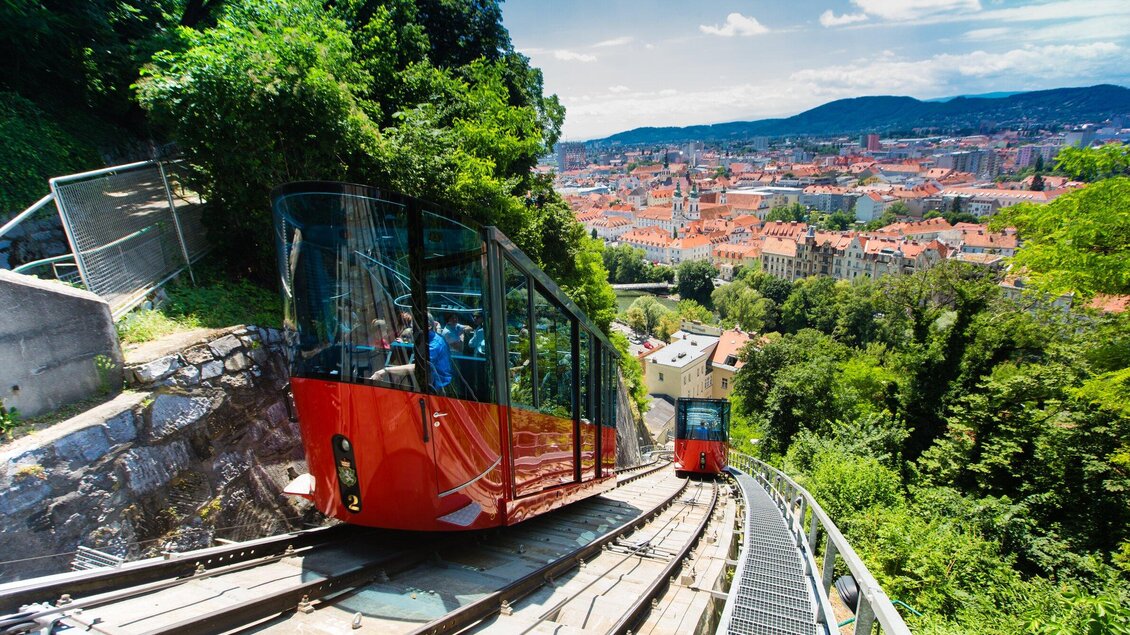 Eine farbige Standseilbahn fährt an einem Hang entlang. Im Hintergrund sieht man eine Stadt mit roten Dächern und einer wunderschönen Landschaft. | © Graz Tourismus - Harry Schiffer