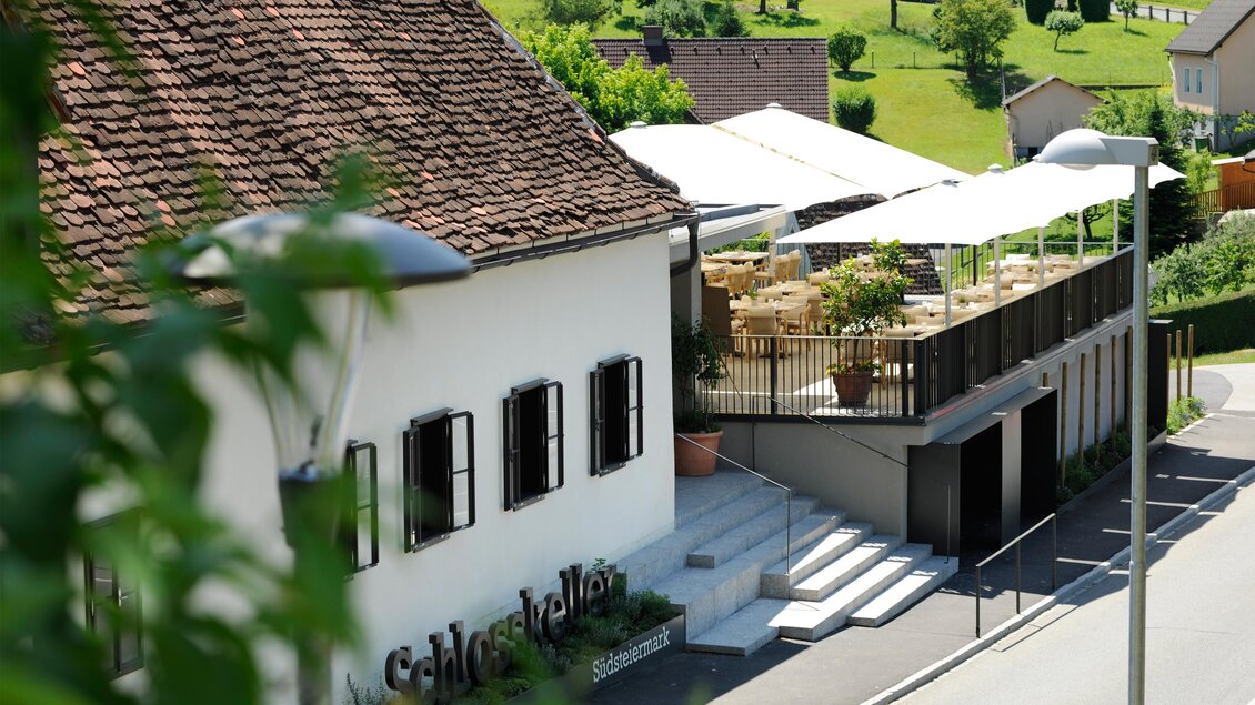 Ein charmantes Gebäude mit einem gemütlichen Außenbereich. Im Hintergrund sieht man grüne Hügel und eine klare, sonnige Umgebung. | © Schlosskeller Südsteiermark