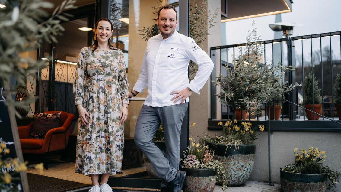 Ein lächelndes Paar steht vor einem Restaurant. Der Mann trägt eine Kochjacke, während die Frau ein blumiges Kleid anhat. | © Hutter