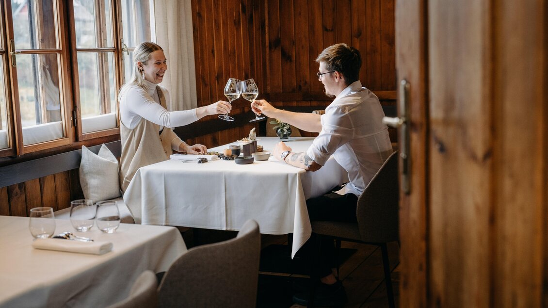 Ein Paar stößt mit Gläsern an und genießt ein romantisches Dinner in einem gemütlichen Holzraum. Der Tisch ist schön gedeckt und das Ambiente ist warm und einladend. | © Hutter