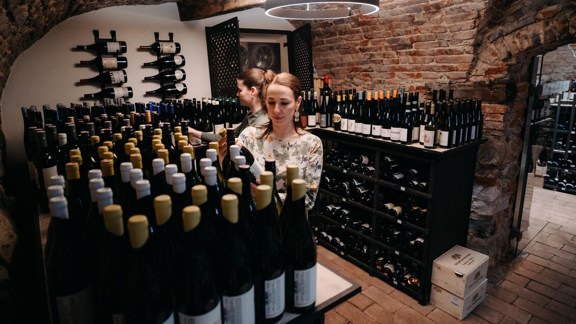 Ein Weinkeller mit vielen Weinflaschen auf Regalen. Zwei Frauen arbeiten im Hintergrund, während sie die Flaschen sortieren. | © Hutter
