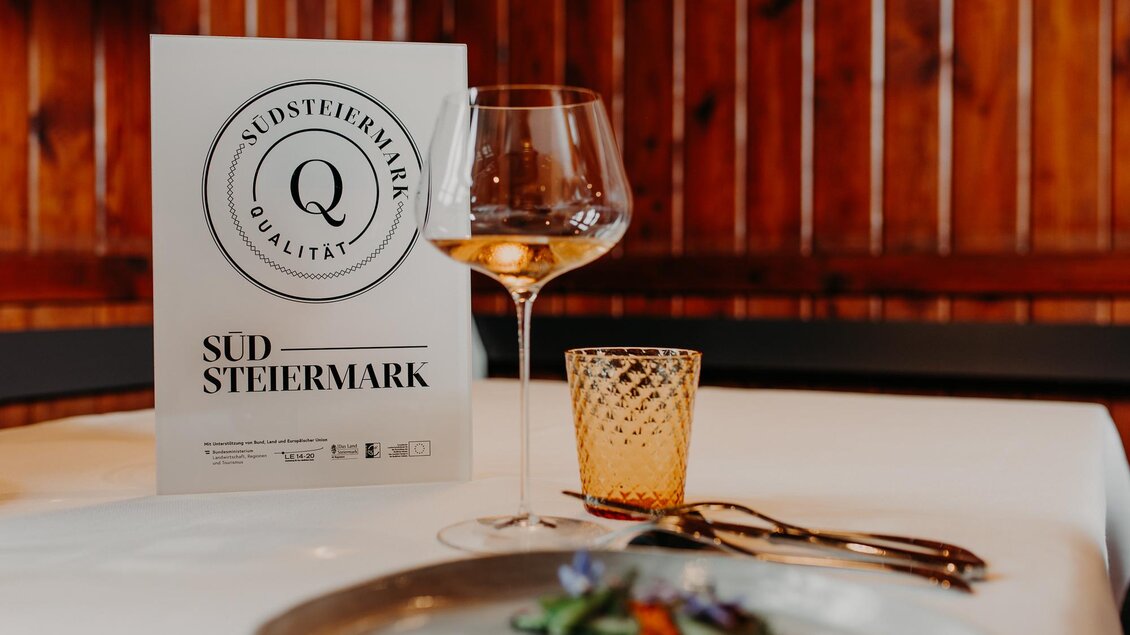 Eine elegannte Tischanordnung mit einem Glas Wein, einer Menükarte und einem Teller mit frischen Zutaten. Die warme Holzumgebung schafft eine gemütliche Atmosphäre. | © © RMSW GmbH/webquartier