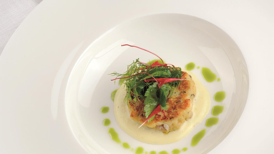 Ein elegant angerichteter Teller mit einem kleinen Küchlein, garniert mit frischen Kräutern. Dazu steht eine weiße Kanne mit grünem Getränk.