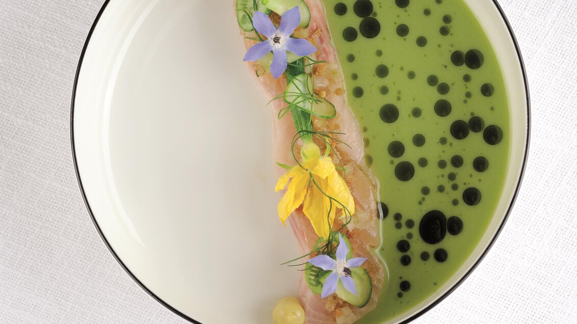 Ein kunstvoll angerichteter Teller mit einer Fischportion, serviert in grünem Fond. Das Gericht ist mit essbaren Blumen und kreativen Saucen dekoriert.