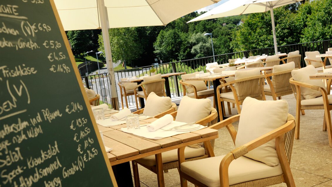 Eine gemütliche Terrasse mit Holzstühlen und einem Speiseplan. Im Hintergrund sind Bäume und Sonnenschirme zu sehen. | © Schlosskeller Südsteiermark