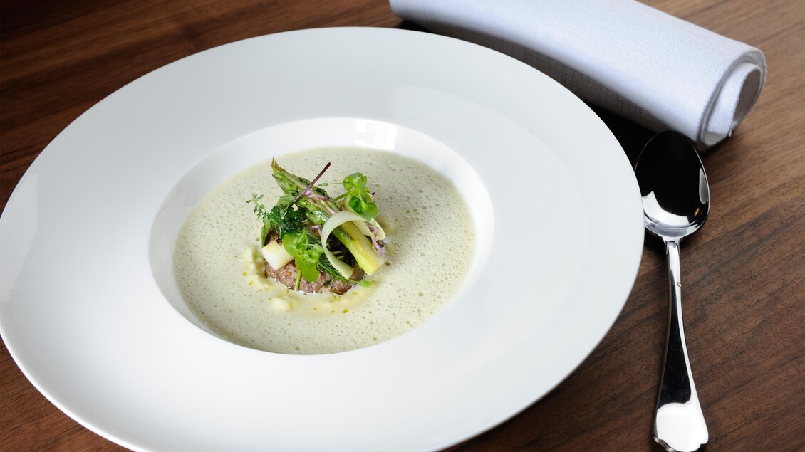 Eine elegante Suppe in einem großen, weißen Teller, garniert mit frischen Kräutern und Gemüse. Neben dem Teller liegt ein Löffel und ein Stoffserviette.