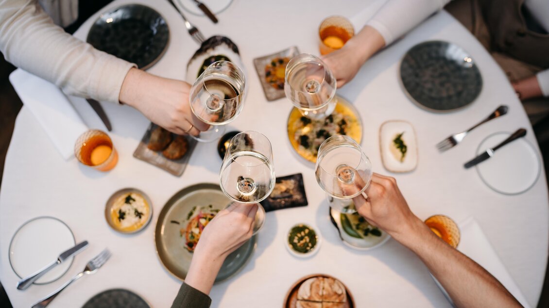 Ein festlich gedeckter Tisch mit verschiedenen Gerichten und Getränken. Mehrere Hände heben Gläser in einem Toast.