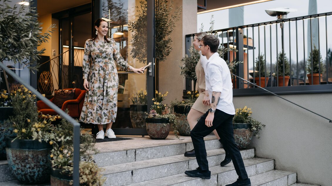 Eine Frau begrüßt zwei Männer, die die Treppe hinaufgehen. Die Szene spielt vor einem modernen Gebäude mit Pflanzen und blühenden Töpfen. | © Hutter