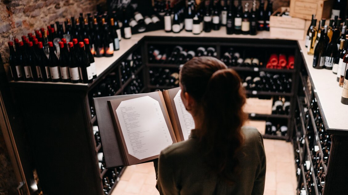 Eine Person steht in einem Weinkeller und betrachtet eine Weinkarte. Um sie herum sind viele Weinflaschen ordentlich aufgereiht. | © Hutter