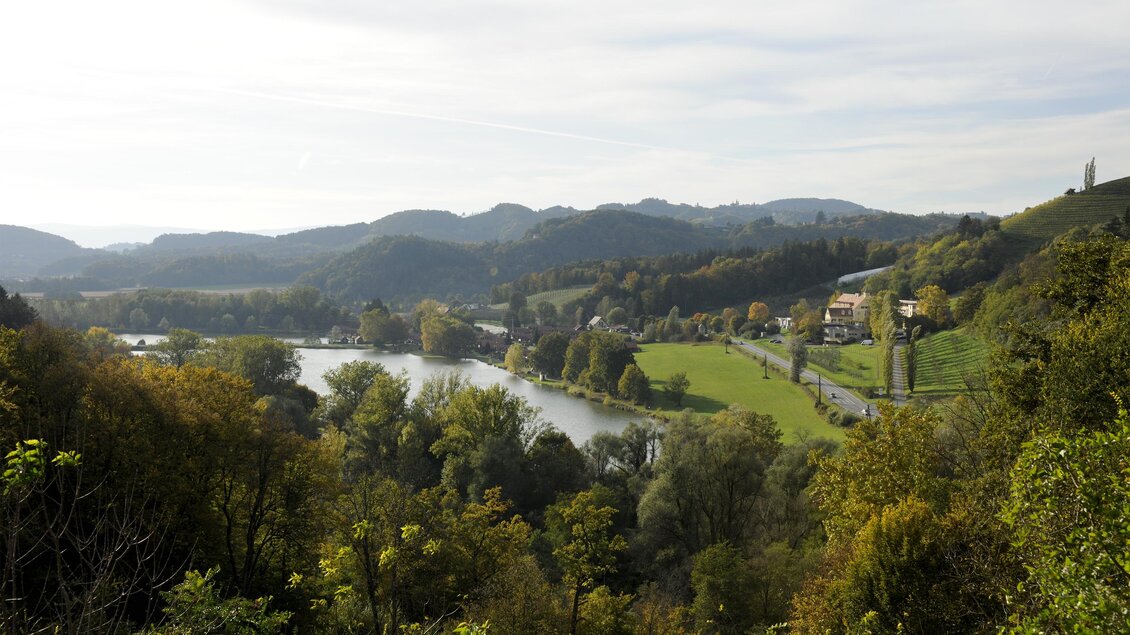 Eine ruhige Landschaft mit einem Fluss, umgeben von Bäumen und sanften Hügeln. Der Himmel ist bewölkt und die Farben der Natur sind lebendig. | © Schlosskeller Südsteiermark