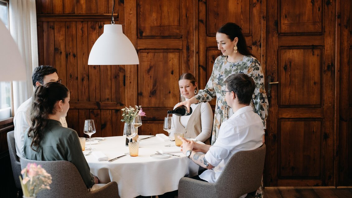 Eine elegante Restaurantatmosphäre mit einem Tisch, der für vier Personen gedeckt ist. Eine Kellnerin serviert Getränke an die Gäste.