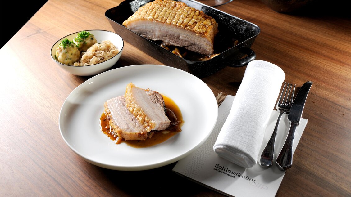 Ein Teller mit zartem Fleisch und knuspriger Kruste, serviert in einer aromatischen Sauce. Daneben eine Schüssel mit Beilagen auf einem Holztisch. | © Schlosskeller Südsteiermark