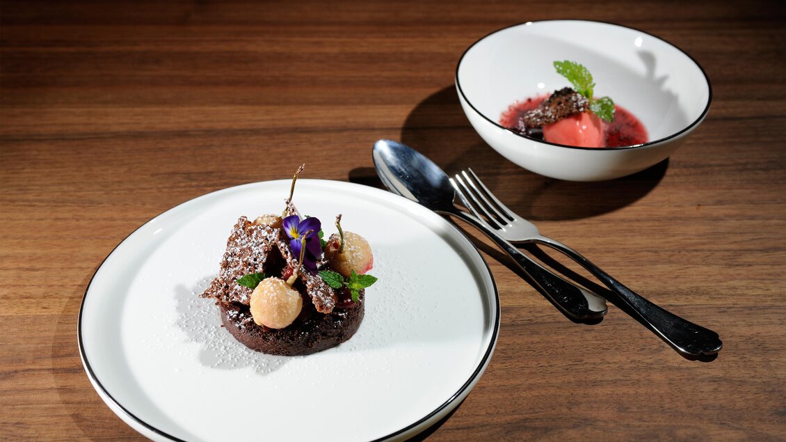 Ein elegant angerichtetes Dessert auf einem weißen Teller, dekoriert mit essbaren Blüten. Nebenan liegt eine Schüssel mit einer erfrischenden Fruchtsoße.