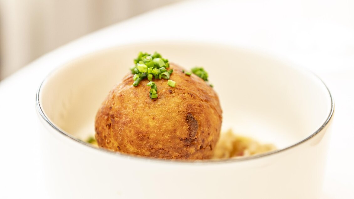 Eine kreative Speise in einer weißen Schüssel, bestehend aus einer frittierten Kugel mit grünen Kräutern obendrauf. Der Hintergrund ist hell und minimalistisch.