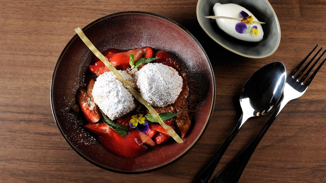 Ein köstliches Dessert mit zwei runden, pudrigen Küchlein auf frischen Erdbeeren. Daneben befindet sich eine kleine Schale mit Eis und essbaren Blumen. | © Schlosskeller Südsteiermark