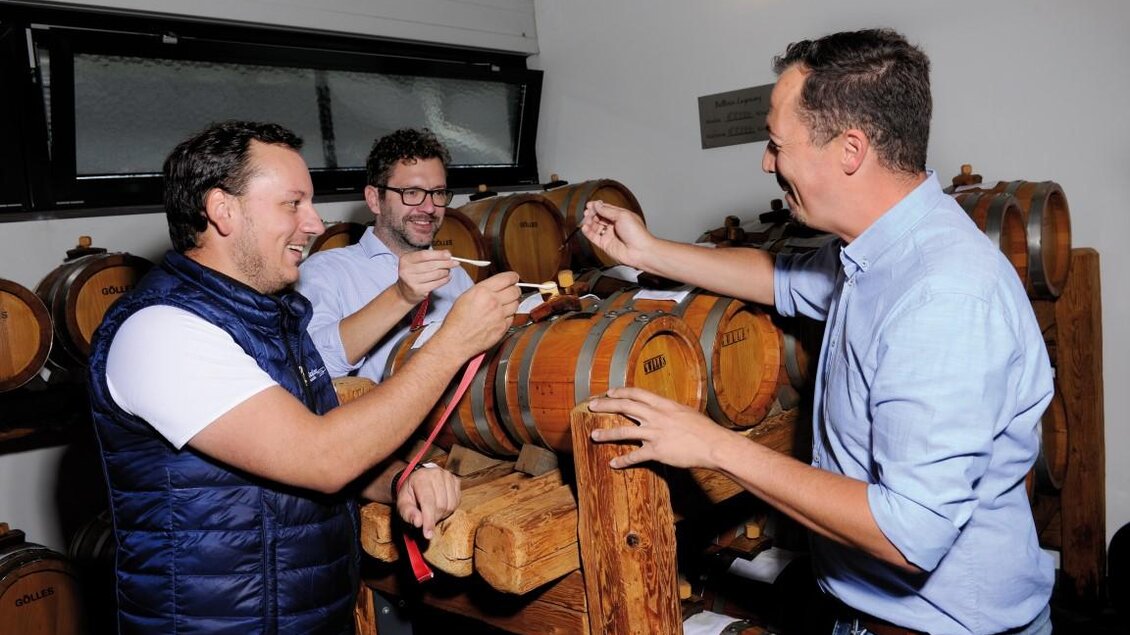 Drei Männer stehen vor Holzfässern und unterhalten sich. Sie scheinen Spaß zu haben und verkosten möglicherweise Wein.