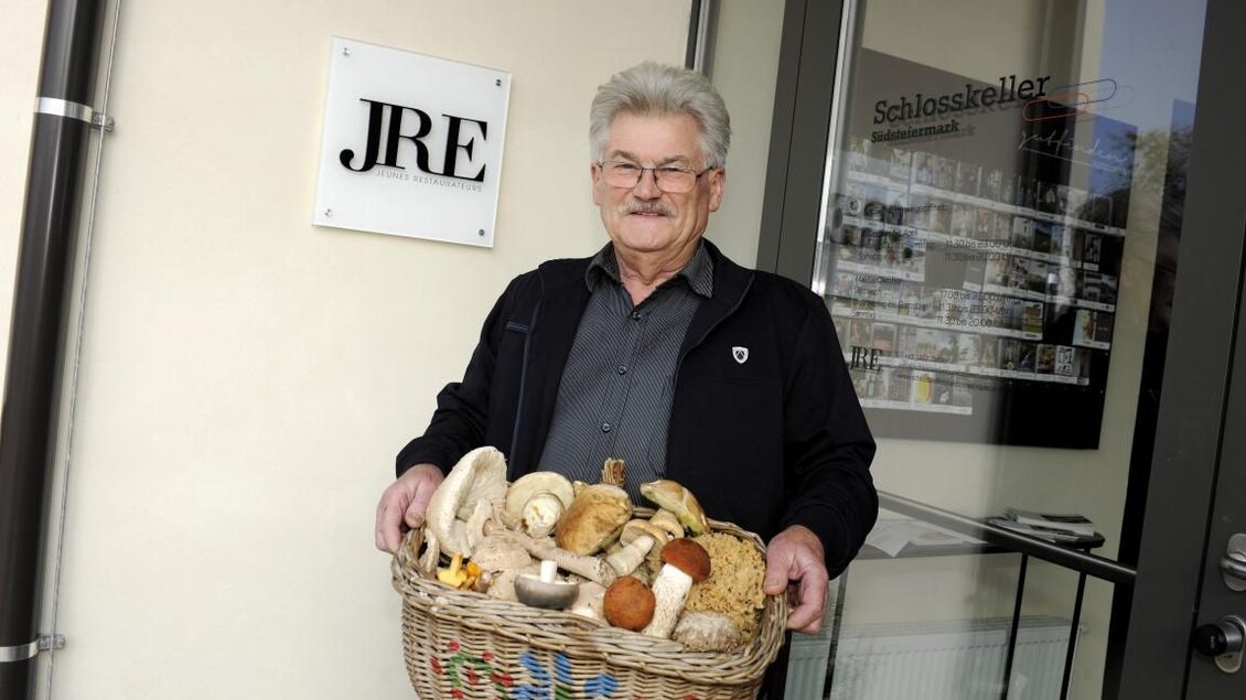 Ein älterer Mann steht vor einem Eingang und hält einen Korb mit verschiedenen Pilzen. Er trägt eine dunkle Jacke und lächelt freundlich in die Kamera.