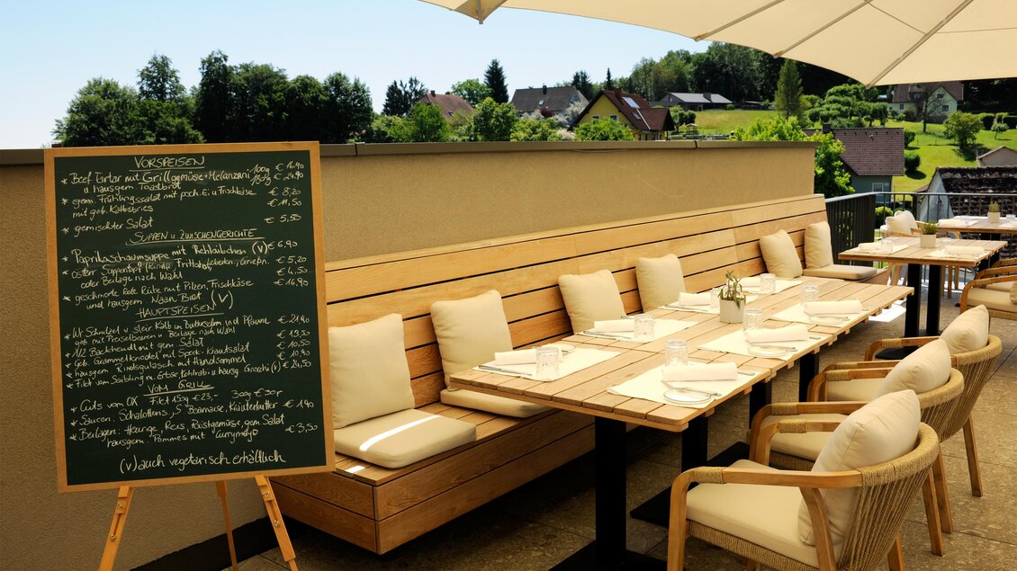 Ein gemütlicher Außenbereich mit Holzbank und Tischen. Im Hintergrund ist eine Tafel mit der Speisekarte und eine schöne Landschaft zu sehen. | © Schlosskeller Südsteiermark