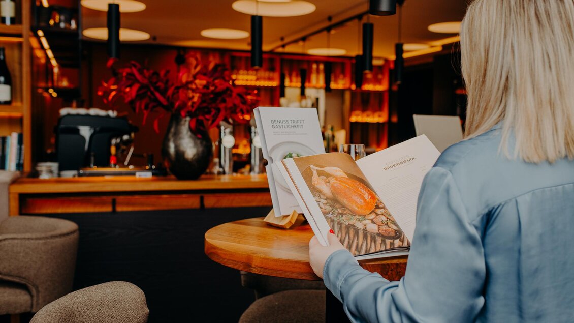 Eine Person sitzt an einem Tisch in einem modernen Café und studiert ein Buch. Im Hintergrund sind ein gut ausgestatteter Barbereich und dekorative Blumen zu sehen. | © © RMSW GmbH/webquartier