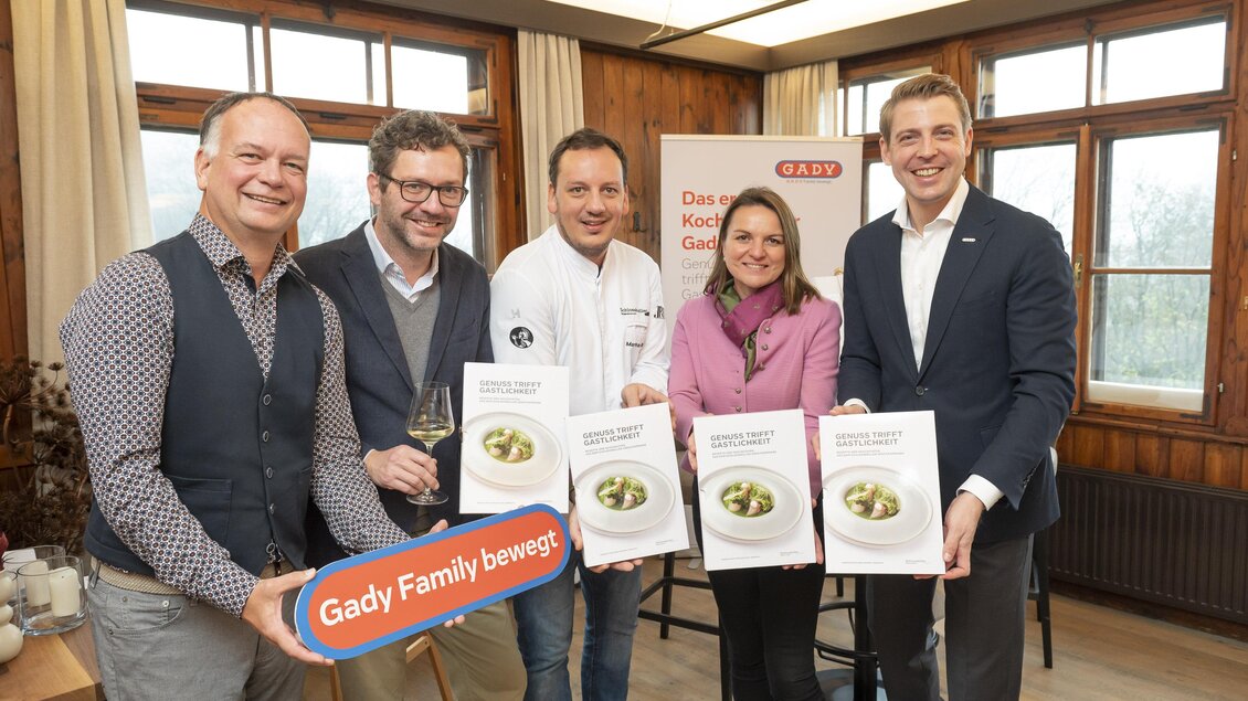 Eine Gruppe von fünf Personen steht zusammen in einem Raum und hält Kochbücher in den Händen. Im Hintergrund sind Fenster zu sehen, die viel Licht hereinlassen.