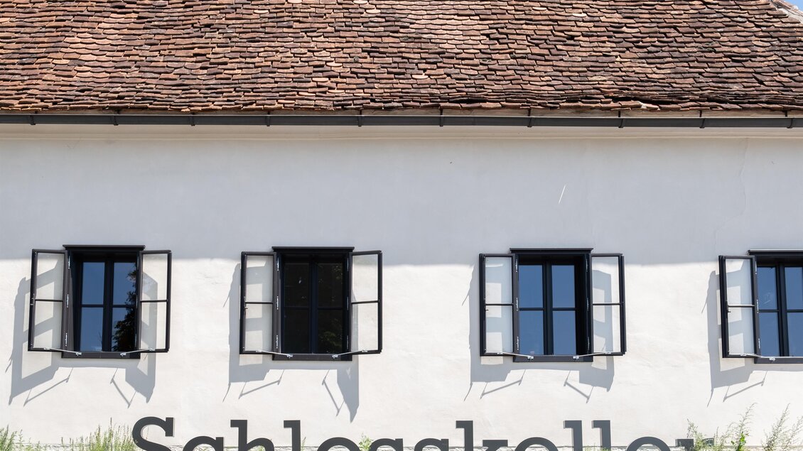 Ein einfaches Gebäude mit einer weißen Wand und einem Satteldach. Auf der Fassade steht der Schriftzug "Schlosskeller" und es gibt mehrere Fenster mit offenen Fensterläden. | © Schlosskeller Südsteiermark