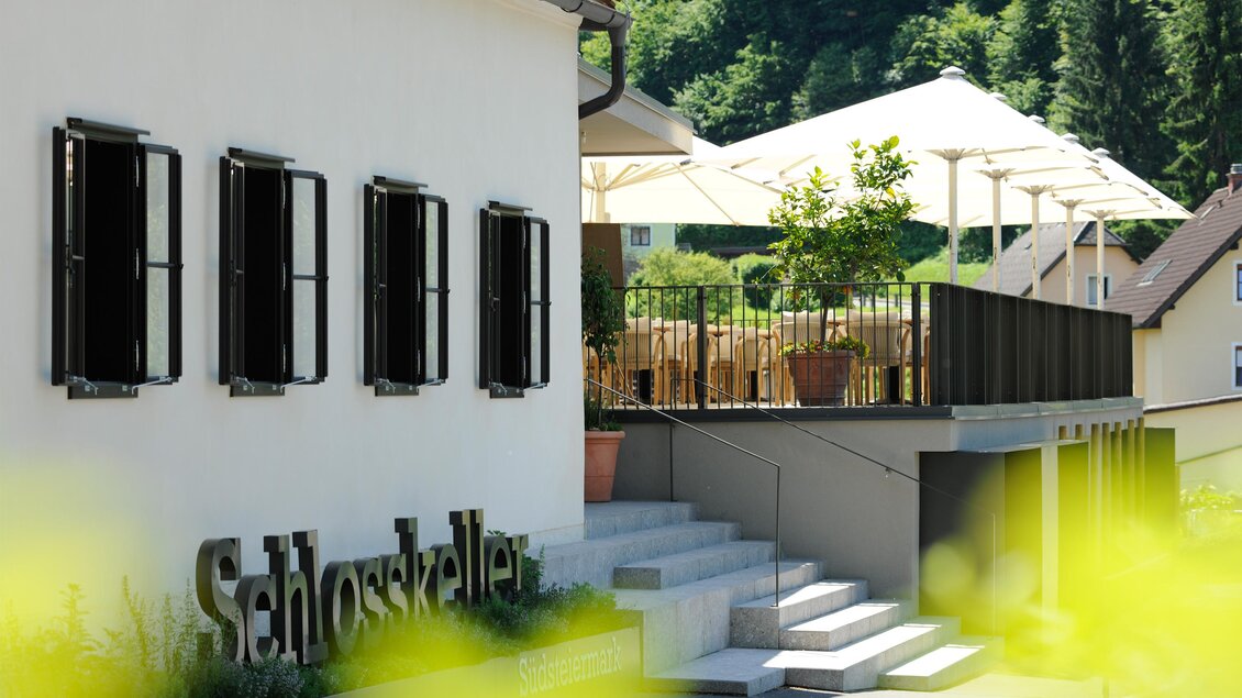 Ein charmantes Gebäude mit Fensterläden und einem einladenden Außenbereich. Sonnenschirme und Pflanzen schaffen eine angenehme Atmosphäre. | © Schlosskeller Südsteiermark