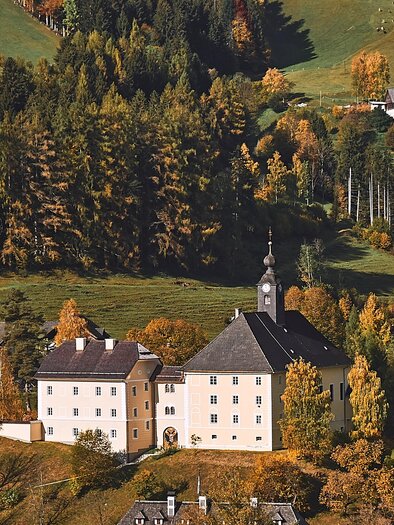 An impressive castle surrounded by colorful trees in autumn. The landscape is diverse and green, with gentle hills and scattered houses. | © Armin Walcher