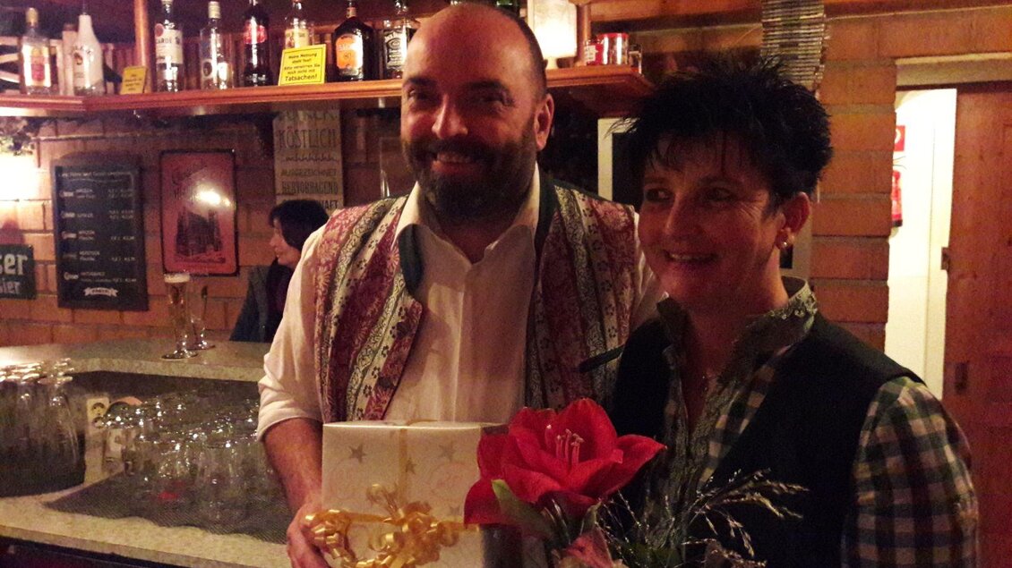Ein Mann und eine Frau stehen lächelnd an der Bar. Sie halten ein Geschenk und einen Blumenstrauß in der Hand. | © Schlosstaverne