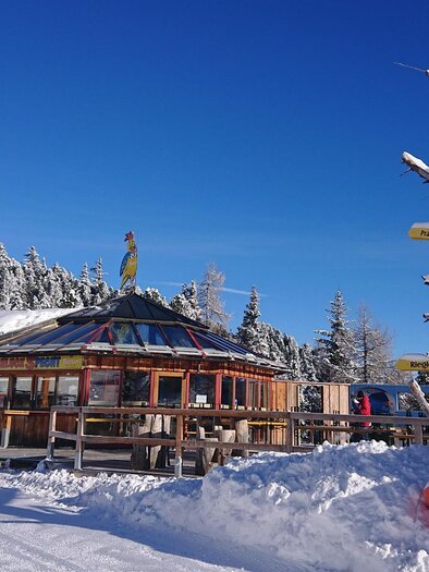 A cozy mountain cabin in the snow under a blue sky. Next to the cabin, there is a tree with signs indicating different directions. | © Schluckspecht