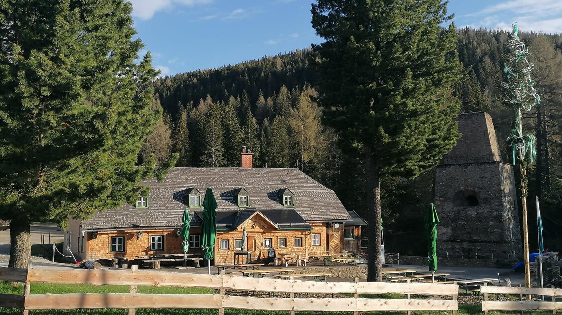 Ein charmantes Holzhaus umgeben von hohen Bäumen und einer idyllischen Landschaft. Im Hintergrund ist ein alter, steinerner Turm zu sehen. | © Herbert Schönhart