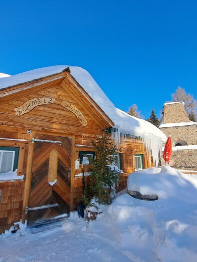 A cozy wooden cabin in the snow, surrounded by snow-covered trees and a clear blue sky. The cabin has a charming, rustic design. | © Claudia Bachler