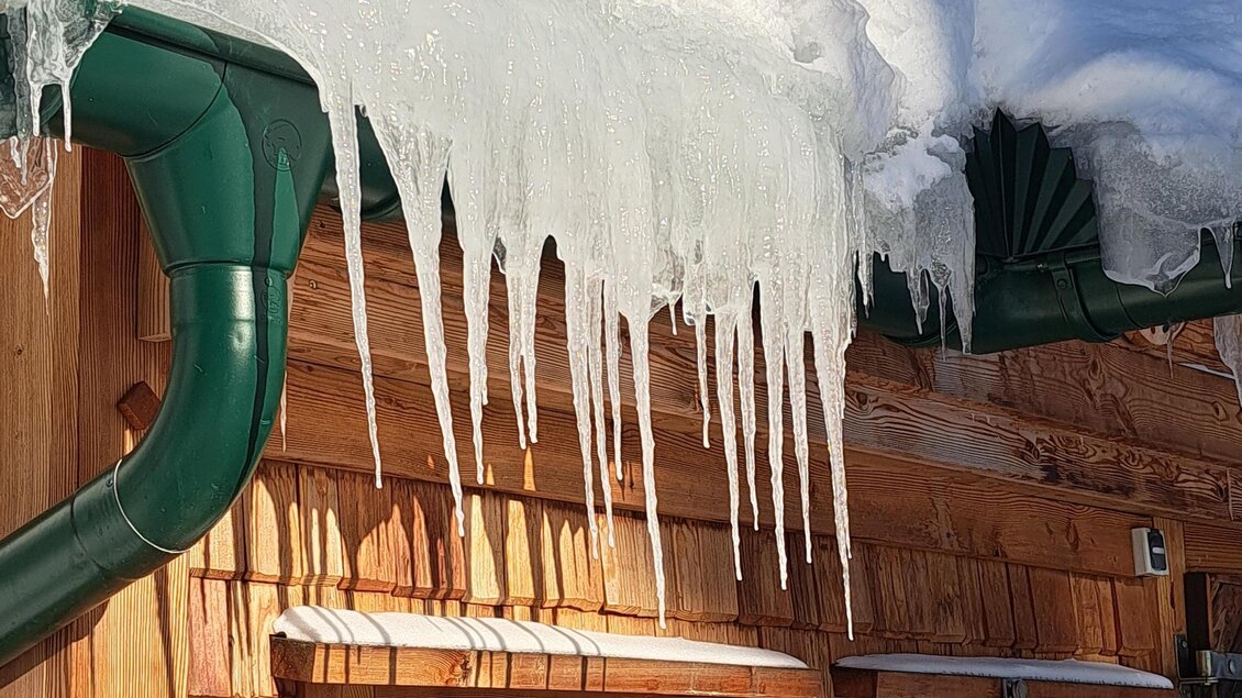 Eiszapfen hängen von einem schneebedeckten Dach. Das Holzhaus hat grüne Fensterrahmen und klare blaue Himmel im Hintergrund. | © Claudia Bachler