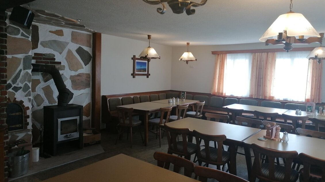 Ein gemütlicher Gastraum mit Holztischen und -stühlen. An der Wand befindet sich ein Kamin, und es gibt große Fenster mit Vorhängen. | © Herbert Schönhart